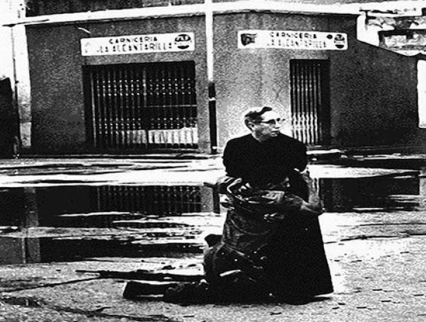 El capellán Luis María Padilla, de la iglesia de Puerto Cabello, parado en el medio de la calle ayudando a un soldado del Gobierno herido durante la revuelta. Esta foto ganó el premio Pulitzer de fotografía de 1963